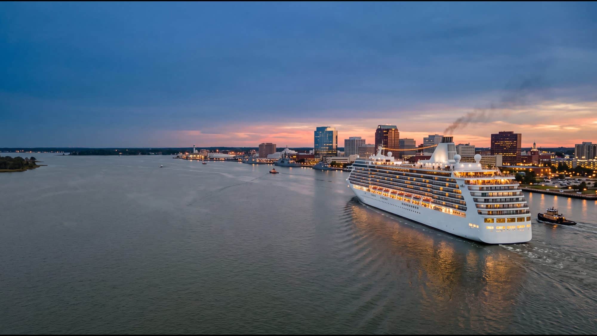 A white cruise ship sailing down the Elizabeth River at dusk with its lights on, the Norfolk skyline behind